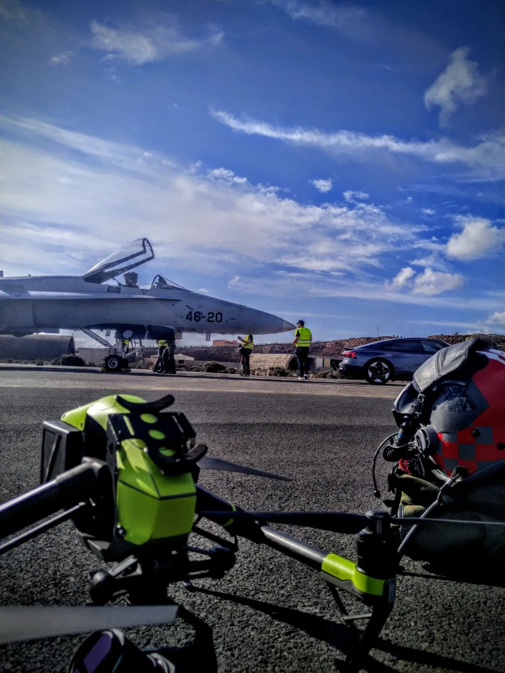 Caza F-18 con la cabina abierta en la plataforma de una base aérea; en primer plano, un dron de cine y unas gafas FPV, y alrededor del avión personal de tierra con chalecos de alta visibilidad