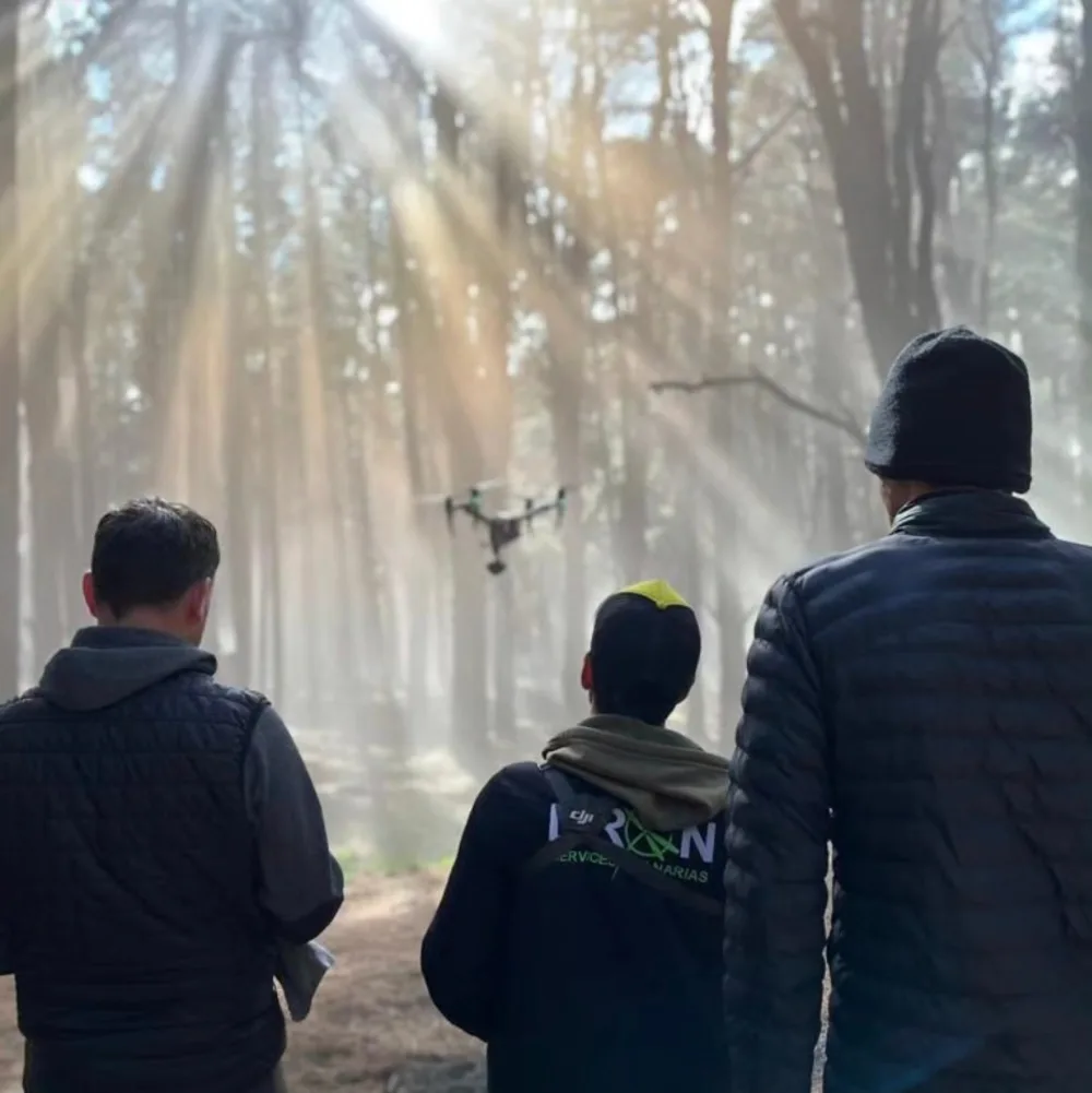 Tres personas de espaldas en un pinar con haces de luz entre los troncos; en el centro, el cinelifter FPV de Dron Services Canarias en vuelo