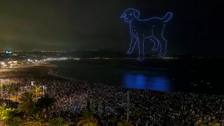 Formación nocturna de drones dibujando la figura de un baifo sobre la bahía, con público en la playa