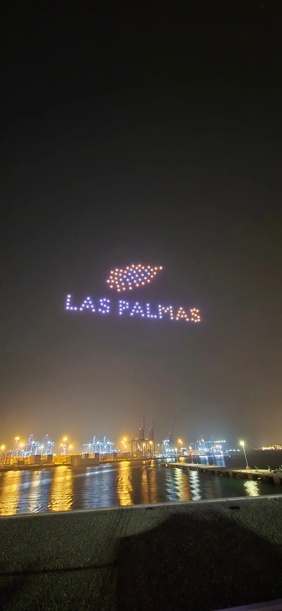 Formación nocturna de drones sobre el Puerto de Las Palmas dibujando el texto «LAS PALMAS» con una figura luminosa encima; al fondo, las grúas del puerto iluminadas