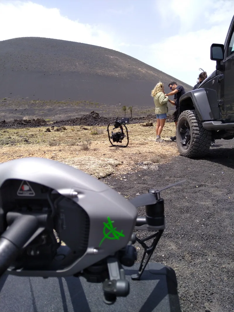 Dron con el logo de Dron Services Canarias en primer plano; al fondo, equipo de rodaje con un estabilizador de cámara en un paisaje volcánico